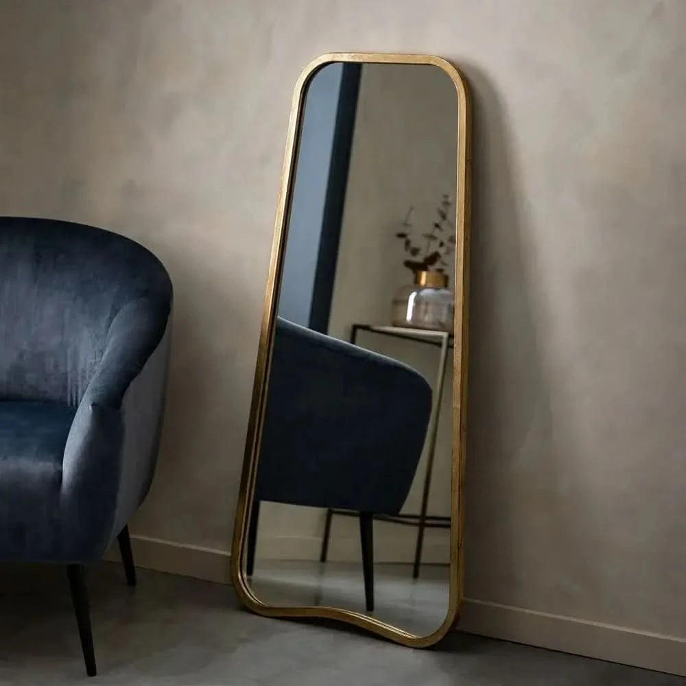 The hollywood mirrors Valentina Gold Full Length Mirror (123cm x 56.5cm) leans against a beige wall, its unique shape reflecting a small table with a vase, while a dark blue velvet chair sits nearby.